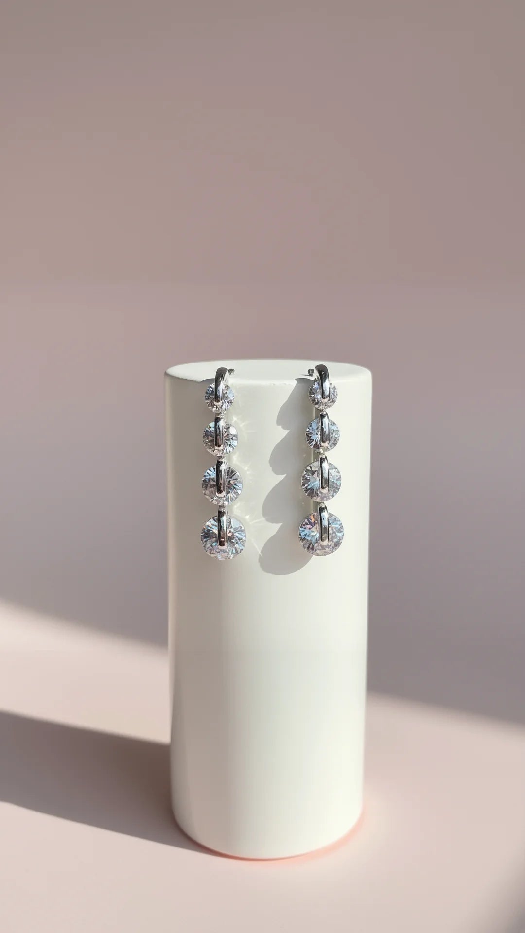 Pair of dangling earrings on a cylindrical white stand against a light pink background
