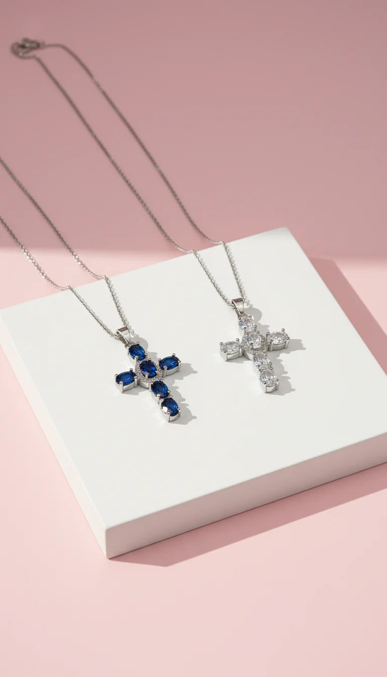 Two silver cross necklaces with blue and clear gemstones on a white box against a pink background.