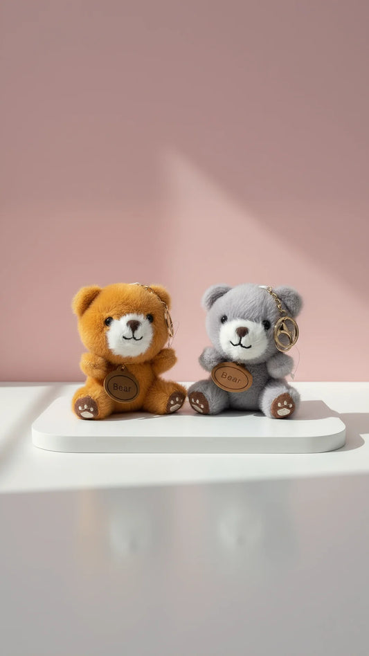 Two decorative teddy bears keychains in brown and gray colors, displayed on a white surface against a pink background