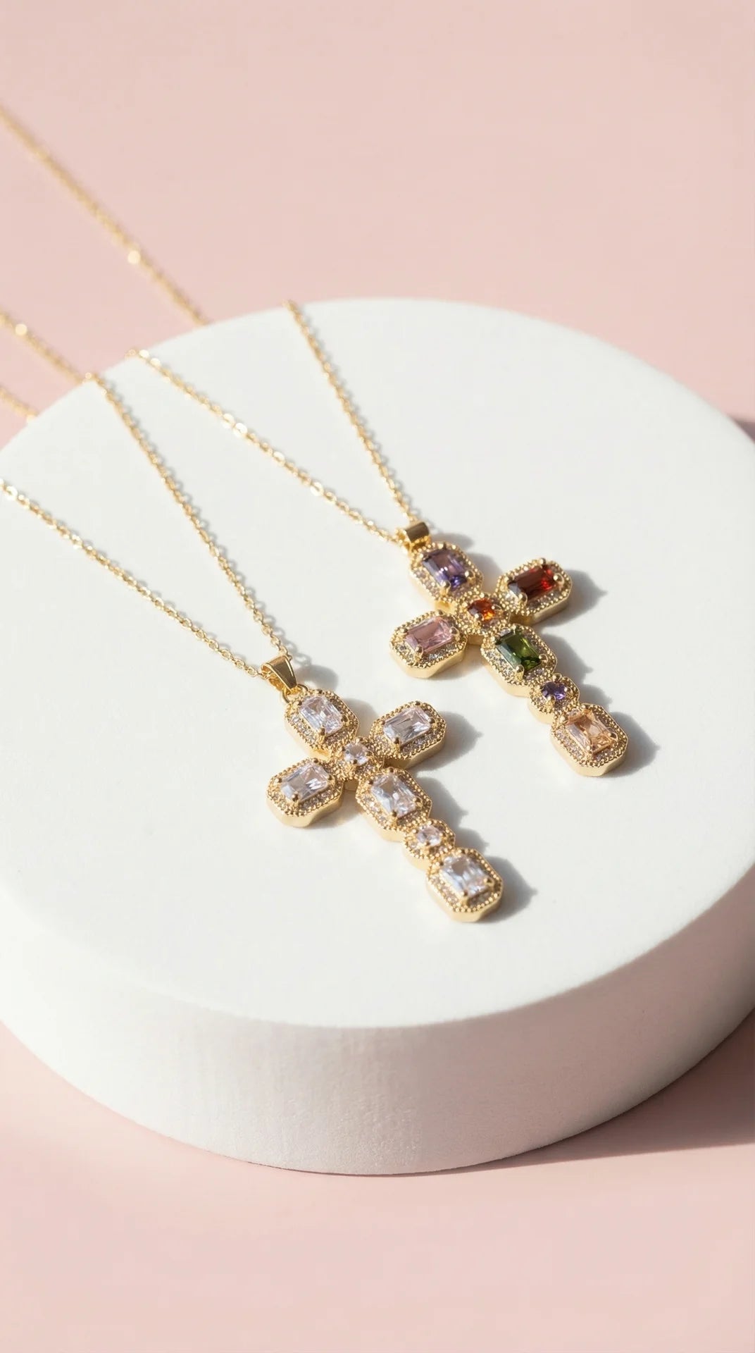 Gold cross necklaces with gemstones on a white stand against a pink background