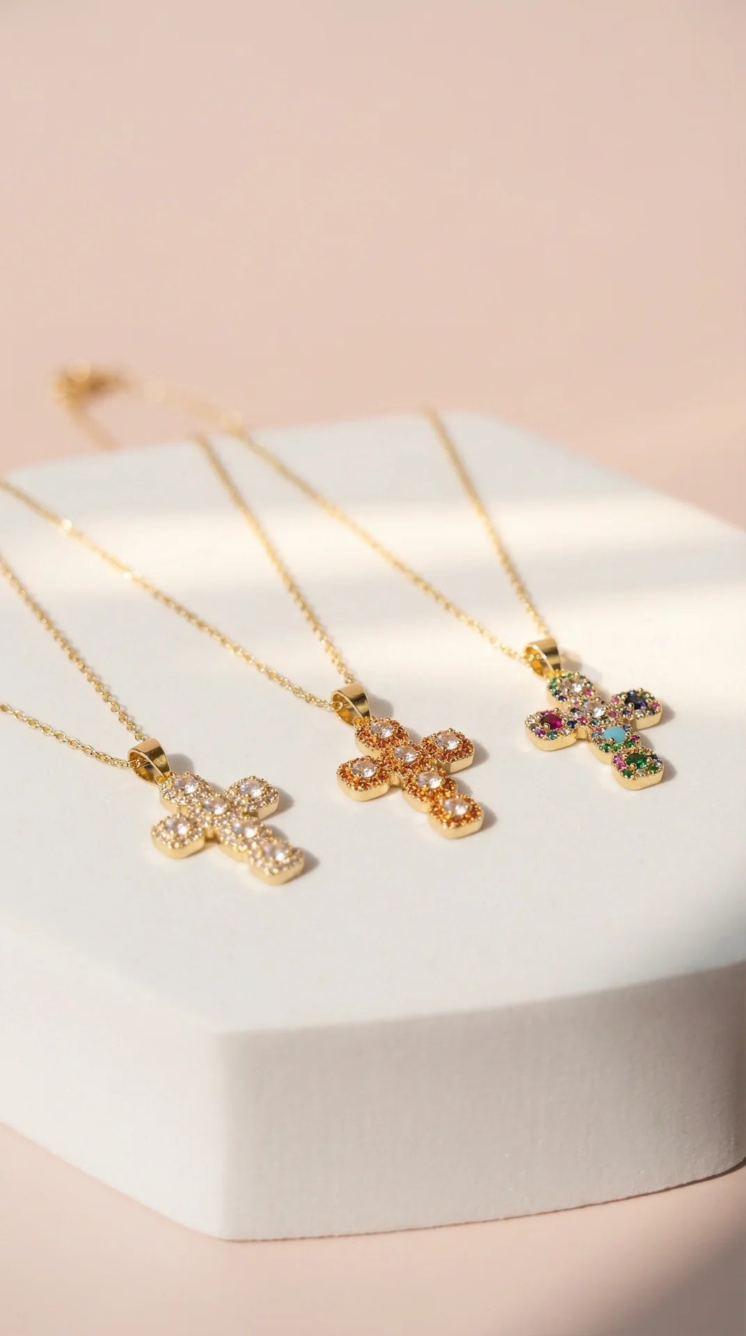 Three gold cross necklaces with colorful gemstones on a white stand against a light pink background.