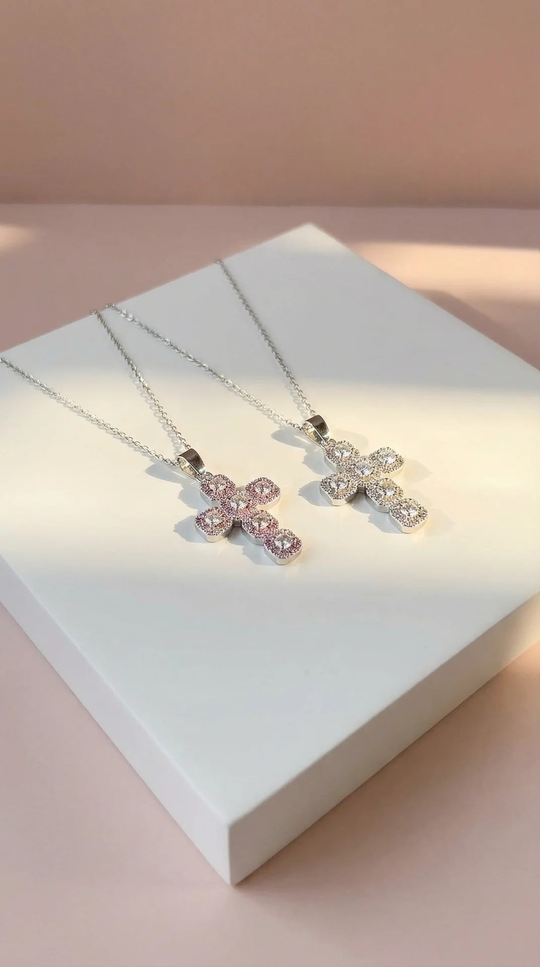 Two silver cross necklaces with pink accents on a white box.