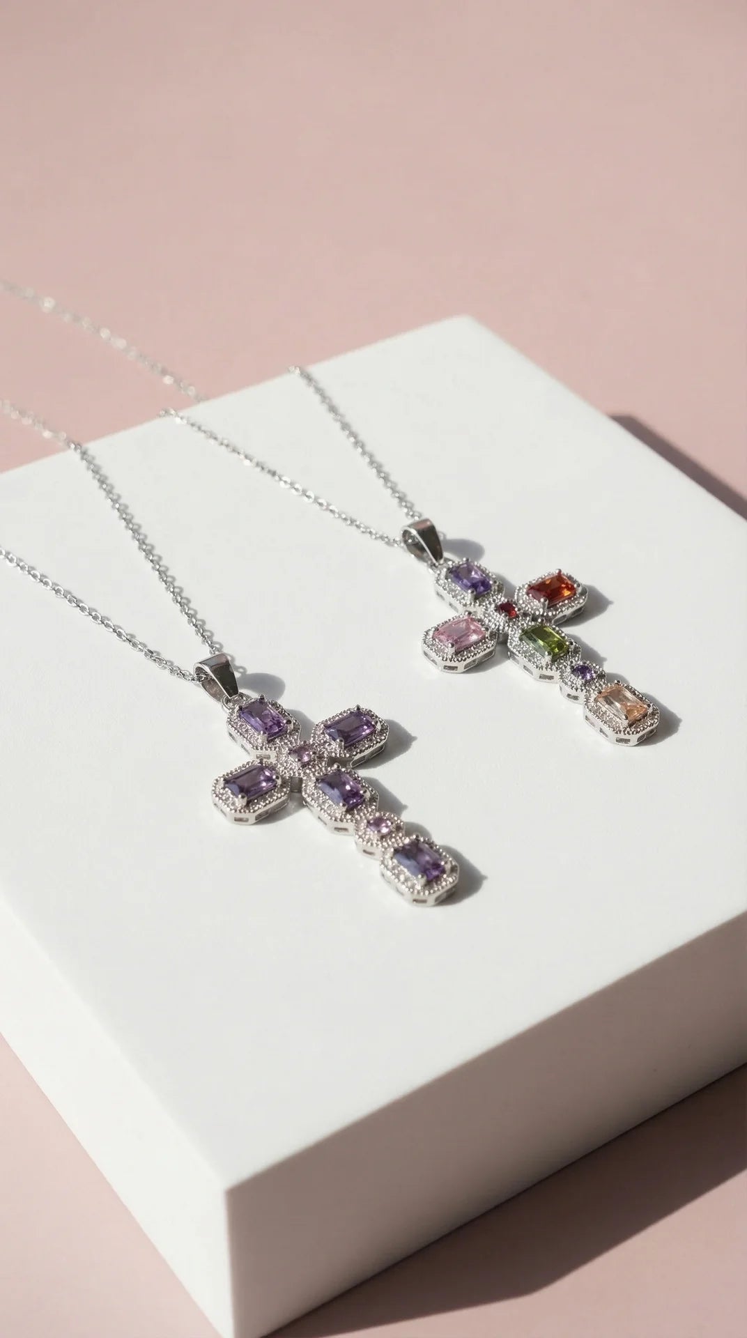 Two cross-shaped necklaces with gemstones on a white box against a pink background
