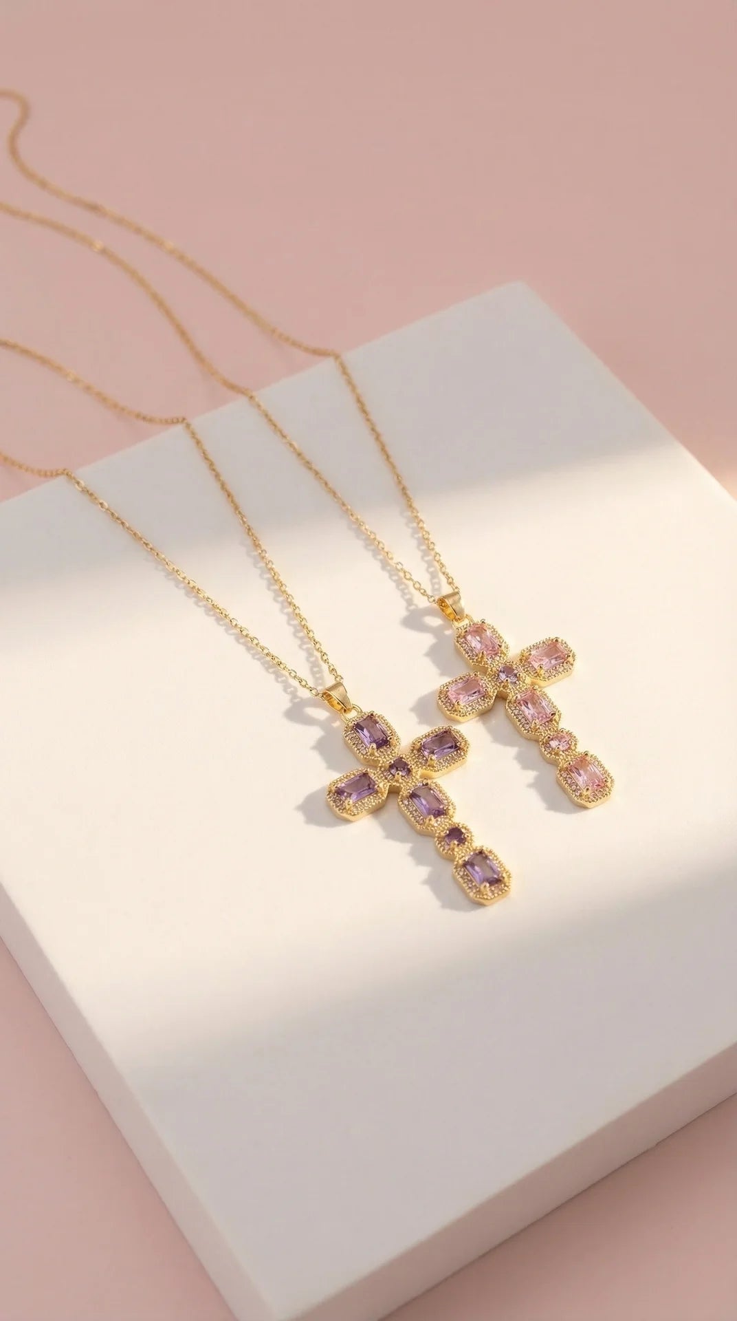 Gold cross necklaces with pink gemstones on a white box with a pink background