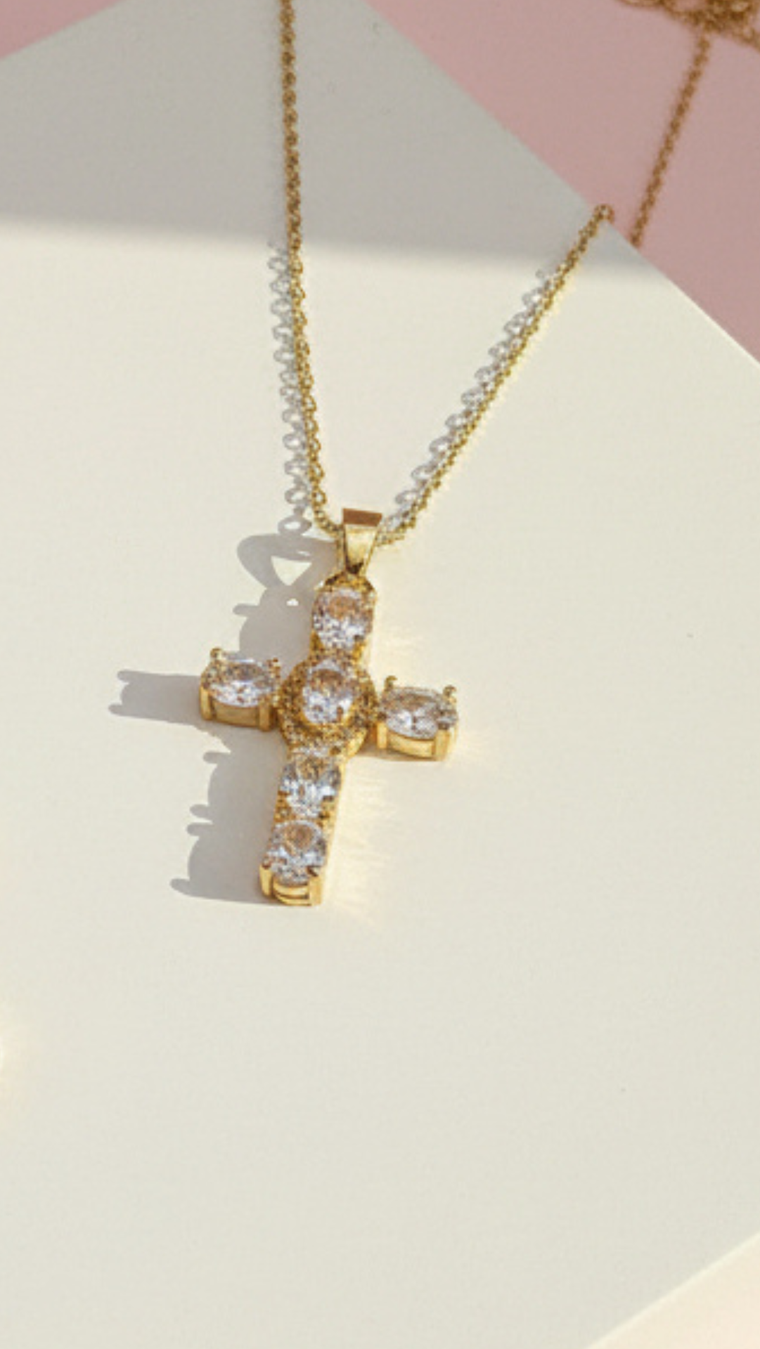 Golden cross pendant with clear crystals displayed on a white surface with pink background
