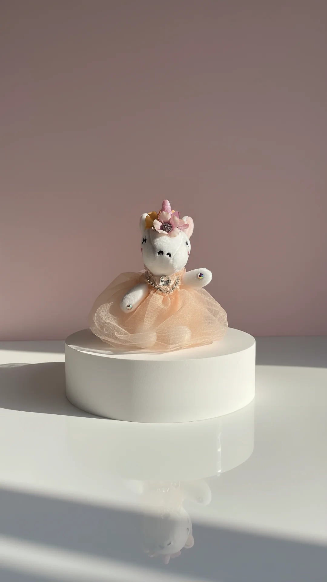 Plush white unicorn keychain with light orange dress, displayed on a white surface against a pink background